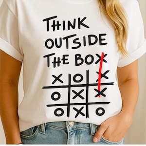 Może przedstawiać: Biała koszulka z czarnym napisem "THINK OUTSIDE THE BOX" nad kratką do gry w kółko i krzyżyk. Czerwona linia przecina kratkę. Koszulka jest noszona z niebieskimi dżinsami.