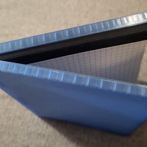 Puede incluir: Un cuaderno de tapa dura azul con cierre de banda elástica negra. El cuaderno está abierto para revelar una página cuadriculada.