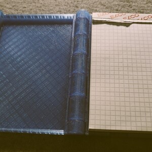 Könnte beinhalten: Ein blaues, 3D-gedrucktes Notizbuch mit einem karierten Papiereinsatz. Das Notizbuch hat einen Rücken, der aus mehreren Schichten blauen Kunststoffs besteht.
