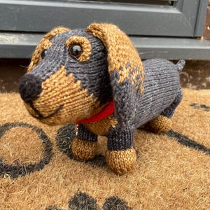 May include: A knitted dachshund toy with brown and gray fur, brown ears, and a red collar. The toy is standing on a brown doormat with paw prints.