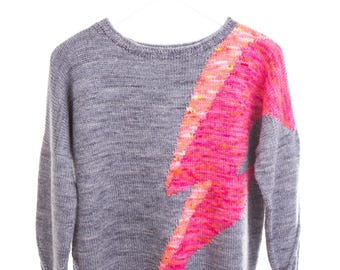May include: A gray knit sweater with a vibrant pink and orange lightning bolt design on the front. The sweater is displayed on a white hanger against a white background. The lightning bolt is a prominent feature.