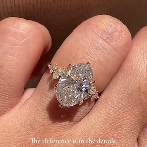 May include: A gold ring featuring a large, oval-cut diamond as the centerpiece, with smaller accent stones on the band. The ring is held up by a hand. Text on the image: "The difference is in the details."