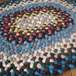 May include: Braided rug with a circular design. The rug features a variety of colors, including blue, gray, yellow, red, and black. The rug is made of woven fabric strips and is suitable for a home decor item.
