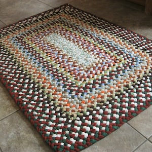 May include: Rectangular braided rug with a mix of colors including red, green, white, brown, orange, and blue. The rug has a border of dark red and green, and the center features lighter shades.