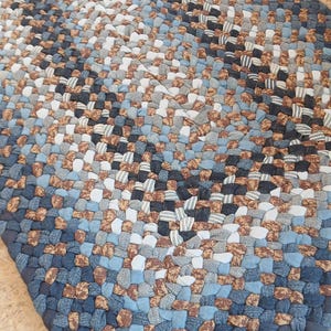 May include: Braided rug in shades of blue, brown, and white. The rug is rectangular and made of woven fabric strips. The color palette includes denim blue, light blue, brown, and white, creating a textured, handcrafted appearance.