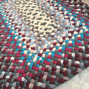 May include: Braided rug with a mix of colors including cream, burgundy, teal, and gray. The rug has a rectangular shape and is made of woven fabric strips. The pattern is a mix of solid and patterned fabrics.