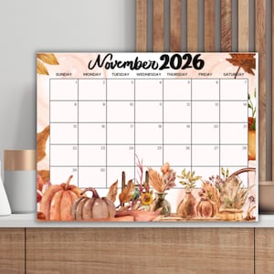 May include: A November 2026 calendar with a watercolour design of pumpkins, autumn leaves, and vases. The calendar grid is on a white background with a peach border. The calendar is displayed on a white shelf, with decorative vases.