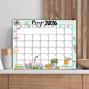 Puede incluir: Calendario decorativo de mayo de 2026 con temática floral. El calendario presenta un fondo verde claro con ilustraciones de acuarela de flores, mariposas y una bicicleta. El calendario se muestra sobre una superficie blanca.