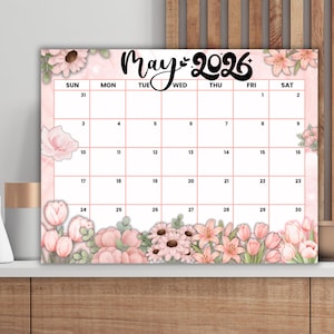Peut inclure: Calendrier de mai 2026 avec un motif floral. Le calendrier présente un fond rose à l'aquarelle avec diverses fleurs, dont des tulipes et des marguerites. Les jours de la semaine sont indiqués en haut, avec le mois et l'année dans une police décorative.
