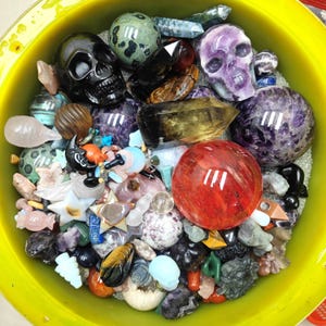 May include: A yellow bowl containing an assortment of polished gemstones and carved items. The collection features crystal skulls, spheres, and figurines in diverse colours, including black, purple, red, and green. The objects are displayed on a bed of small, shiny granules.