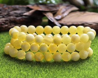 May include: Three strands of yellow beaded bracelets are displayed on a green surface. The beads are round and have a smooth texture. The background includes greenery and a piece of wood.