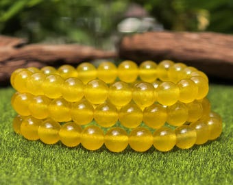 May include: Three yellow beaded bracelets stacked on top of each other. The bracelets are made of round, polished beads. The background is a blurred green with a piece of wood.