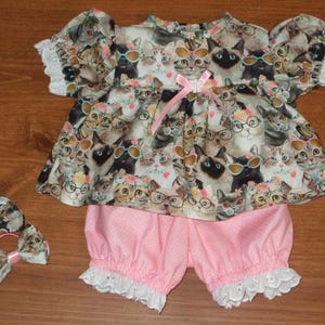 May include: A baby outfit featuring a top and bloomers, along with two matching hair bows. The top is patterned with cats wearing glasses, and the bloomers are pink with white polka dots. The outfit has white lace trim.