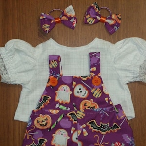 May include: A white short-sleeved blouse with lace-trimmed sleeves, a purple overall-style garment featuring a Halloween print of ghosts, pumpkins, bats, and candy, and two matching purple hair bows with orange centers.
