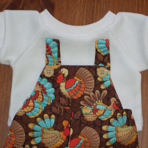 May include: A white short-sleeved shirt paired with brown overalls featuring a repeating pattern of turkeys with teal, orange, and yellow feathers. The overalls have two light-colored buttons at the top of the straps on the front chest.