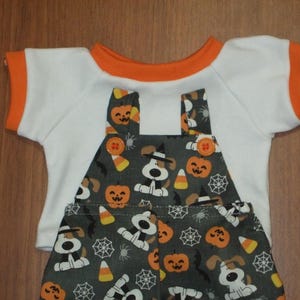 May include: A Halloween-themed outfit for a doll, featuring a white t-shirt with orange trim and a pair of overalls. The overalls are patterned with cartoon dogs, pumpkins, spiderwebs, and candy corn. The outfit is designed for a small doll.