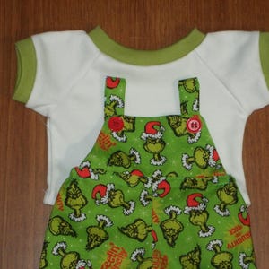May include: A two-piece outfit featuring a white short-sleeved shirt with green trim and a green overall. The overall is patterned with a cartoon character wearing a Santa hat and the words "Feelin' Naughty".