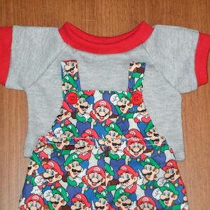 Peut inclure: Un t-shirt gris à manches courtes avec un col rond rouge et des bords de manches rouges, superposé sous une salopette à motif imprimé de Mario et Luigi avec deux petits boutons rouges sur les bretelles avant.