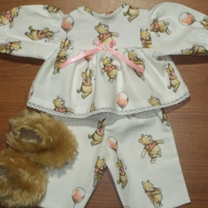 May include: A doll's pajama set featuring a white top and pants with a Winnie the Pooh pattern. The top has lace trim and a pink ribbon. Matching brown fuzzy slippers complete the set. The pajamas are designed for a doll.