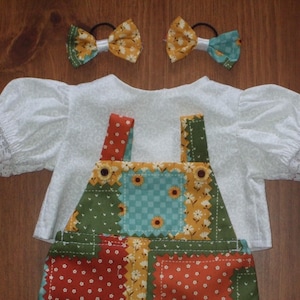 May include: A white short-sleeved blouse with lace-trimmed cuffs, a pair of patchwork overalls featuring green, yellow, and red floral patterns, and two matching hair bows with yellow and teal fabric, all displayed as a coordinated set.
