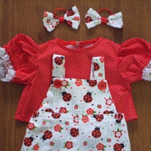 May include: A red and white polka dot shirt with lace trim on the sleeves and a white and red ladybug print overall with red buttons. Two red and white ladybug print hair bows are included.