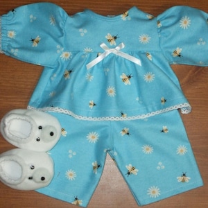 Pode incluir: Um conjunto de pijama para boneca bebé em azul claro, com um padrão de margaridas e abelhas. O conjunto inclui um top com mangas bufantes e um laço branco, calções a condizer e botinhas brancas com olhos pretos. As bordas são aparadas com renda branca.