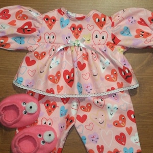 May include: A pink doll pajama set with a heart pattern, featuring a top with puff sleeves and lace trim, matching pants, and pink slippers with googly eyes. The set includes a white ribbon and a small flower.