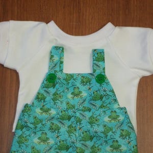 Puede incluir: Una camiseta blanca de manga corta con un peto turquesa con estampado de ranas. El peto tiene botones y tirantes verdes. El estampado de ranas incluye varias poses de ranas y plantas acuáticas. El conjunto está diseñado para un bebé o un niño pequeño.