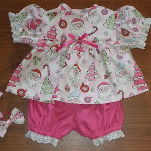 May include: A short-sleeved top and bloomer set featuring a pink and white checkered pattern with Christmas motifs including Santa Claus, trees, and ornaments. The top has a pink ribbon bow at the neckline and white eyelet lace trim.