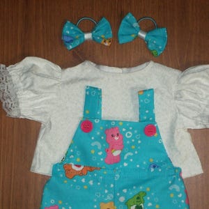 May include: A doll outfit featuring a white top with lace trim and a blue Care Bear-themed overall. Two blue hair bows with white accents complete the set. The overall has pink buttons.