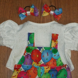 May include: A white top with lace trim sleeves and a colourful balloon print overall. Two matching hair bows with a similar balloon pattern are also included. The outfit is designed for a doll.