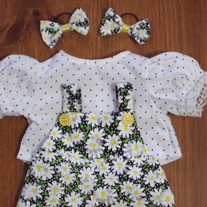 Pode incluir: Um conjunto de boneca com uma parte de cima branca com bolinhas pretas e acabamento em renda, combinada com um macacão com estampa de margaridas. Dois laços de cabelo combinando com estampa de margaridas e detalhes amarelos completam o conjunto. O conjunto tem botões amarelos.