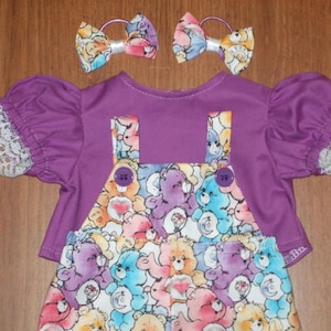 May include: A purple doll outfit with a Care Bears print on the overalls and matching hair bows. The top has short, puffed sleeves with white lace trim. The overalls have purple buttons.