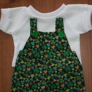 May include: A white short-sleeved shirt paired with a black overall featuring a green, yellow, and blue shamrock pattern. The overall has green buttons on the straps. This outfit is suitable for a baby or toddler.
