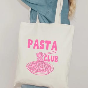 Peut inclure: Sac fourre-tout crème avec les mots "PASTA CLUB" en rose vif. En dessous, une illustration rose d'un bol de spaghettis avec une fourchette. Le sac a deux sangles crème.