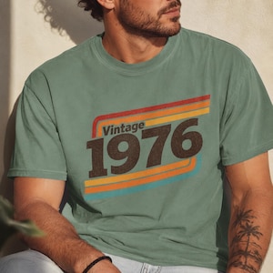 May include: A sage green short-sleeve crew neck t-shirt featuring a graphic print with the text "Vintage 1976" in dark brown. The design includes horizontal stripes in red, orange, yellow, and light blue, creating a retro-inspired aesthetic.