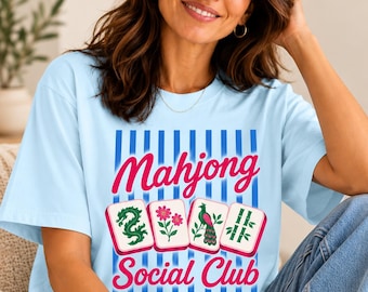Puede incluir: Una camiseta azul claro con un estampado gráfico de rayas verticales azules y el texto "Mahjong Social Club" en rosa. El diseño incluye cuatro fichas de mahjong que muestran un dragón verde, una flor rosa, un pavo real y bambú.