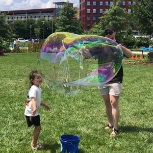 May include: A child watches in awe as a large, iridescent bubble floats in the air. An adult holds the bubble wand, creating the giant bubble. A blue bucket sits on the green grass.