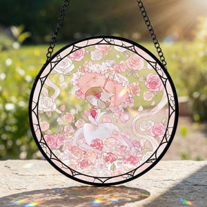 Może przedstawiać: Okrągłe okno witrażowe z czarną metalową ramą i łańcuchem. Grafika przedstawia postać z różowym parasolem i sukienką, otoczoną różowymi różami. Tło jest miękkie, rozmyte zielone.