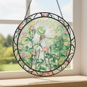 Może przedstawiać: Okrągłe witrażowe dzieło sztuki przedstawiające zielono-białą postać w ogrodzie różowych, żółtych i brzoskwiniowych róż. Grafika jest oprawiona w ciemnobrązową ramę i wisi na łańcuchu.