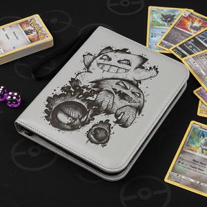 Puede incluir: Un archivador de tarjetas gris claro con una ilustración en blanco y negro de personajes de dibujos animados. El archivador tiene una correa de muñeca negra. Se ven cartas coleccionables y dados morados.
