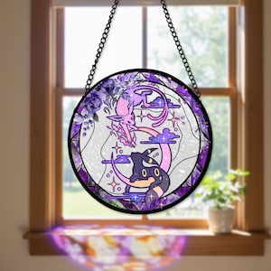 Puede incluir: Vidriera redonda que representa dos personajes de dibujos animados, uno rosa y otro negro, sobre medias lunas. El diseño incluye flores moradas, estrellas y nubes, todo enmarcado en un borde de metal negro. La ventana cuelga de una cadena negra.