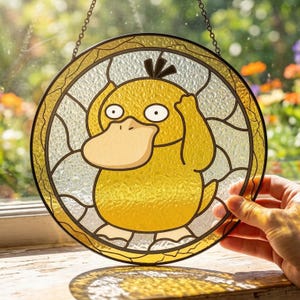 Puede incluir: Una vidriera circular con un pato amarillo de dibujos animados con un pico grande y ojos negros. El pato está dentro de un marco dorado, con una cadena para colgar. El fondo es una mezcla de vidrio transparente y texturizado.