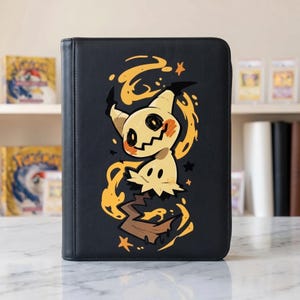 Puede incluir: Carpeta de cartas negra con un diseño de dibujos animados de Mimikyu con detalles en naranja y amarillo. El personaje de Mimikyu tiene un cuerpo color crema y una apariencia dentada y rota. La carpeta está cerrada y tiene una textura suave similar al cuero.