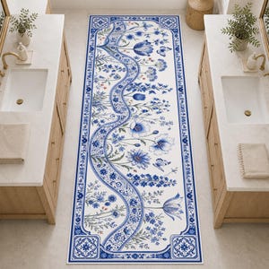 May include: A rectangular white and blue floral patterned rug. The rug features a central flowing design with blue flowers and foliage, framed by a blue border with decorative elements. The rug is designed for a bathroom.