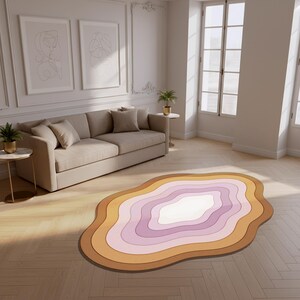 Può includere: Un tappeto astratto a strati nei toni del marrone, arancione, rosa e bianco. Il tappeto ha una forma organica e si trova su un pavimento in parquet chiaro. Un divano beige e opere d'arte incorniciate sono sullo sfondo. La stanza ha una palette di colori neutri.