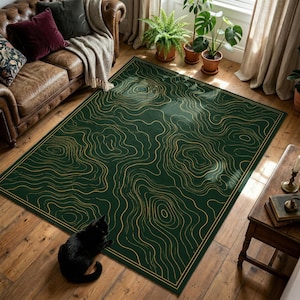 May include: A rectangular green rug with a gold topographic design. The rug is placed on a wooden floor, with a black cat in the lower left corner. A brown leather sofa and plants are in the background.