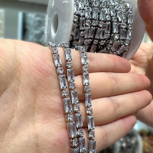 Puede incluir: Primer plano de una brillante cadena plateada con cristales transparentes rectangulares y redondos. La cadena se muestra en una mano, con un carrete de la misma cadena en el fondo. Los cristales reflejan la luz.