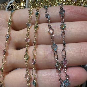 以下が含まれることがあります： 繊細なネックレスのクローズアップ。チェーンは金と銀で、ピンク、青、緑、黄色の色合いのカラフルな宝石で飾られています。宝石はベゼルセッティングでセットされています。ネックレスはニュートラルな背景に表示されています。