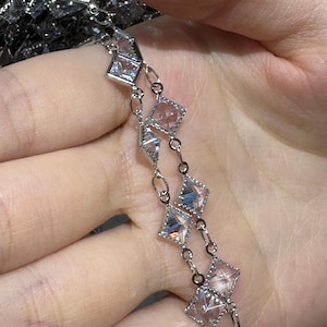 Puede incluir: Una pulsera de plata con un diseño de cadena delicada. La pulsera está adornada con múltiples piedras preciosas claras, de corte cuadrado, cada una engastada en un marco cuadrado. Las piedras preciosas reflejan la luz, creando un efecto brillante.